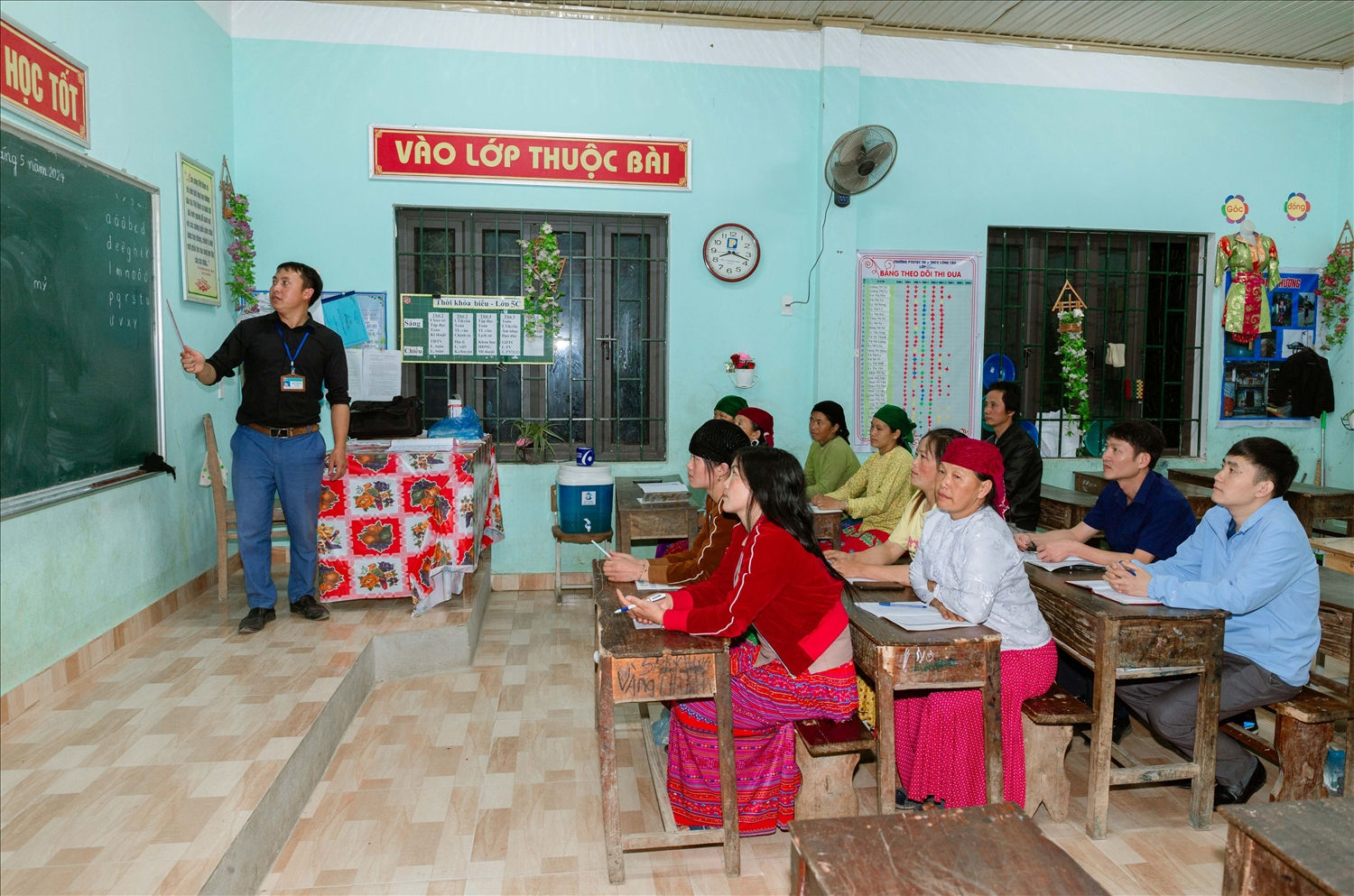 Trên bục giảng, thầy Pó miệt mài dạy chữ cái. Phía dưới, học viên cặm cụi trên trang vở ô ly