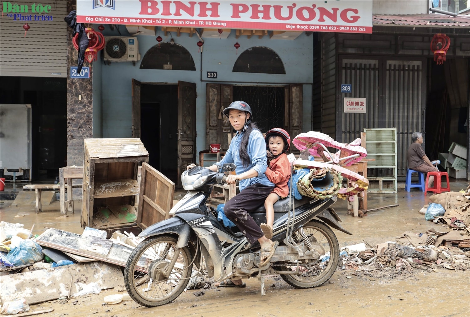 Người mẹ chở con trên chiếc xe máy lấm lem bùn đất, mang theo những đồ dùng còn gom góp lại sau lũ.